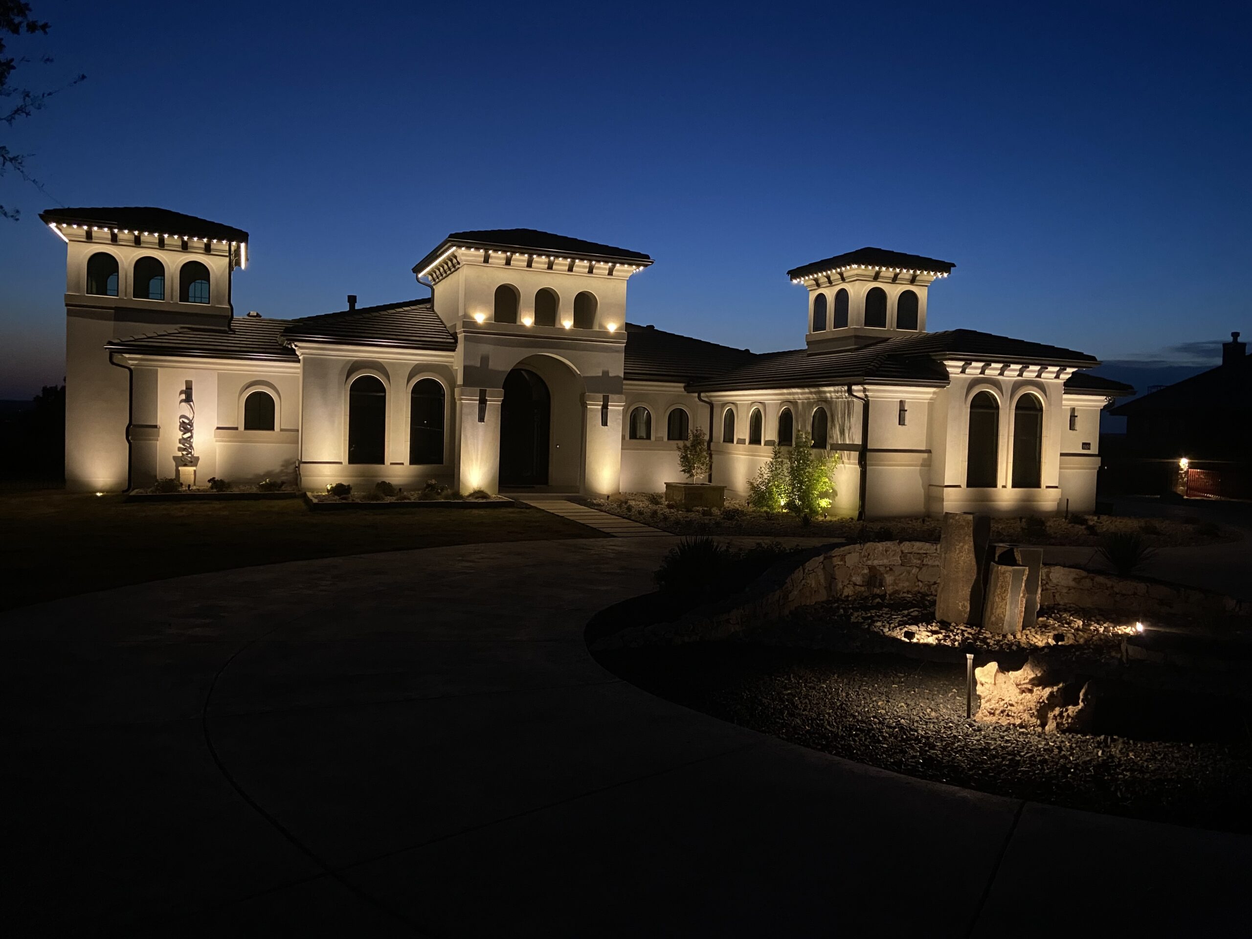 A house illuminated by Astoria Lighting at dusk. The lighting highlights the architectural details of the house, including arched windows and entryways, and the textured facade.
