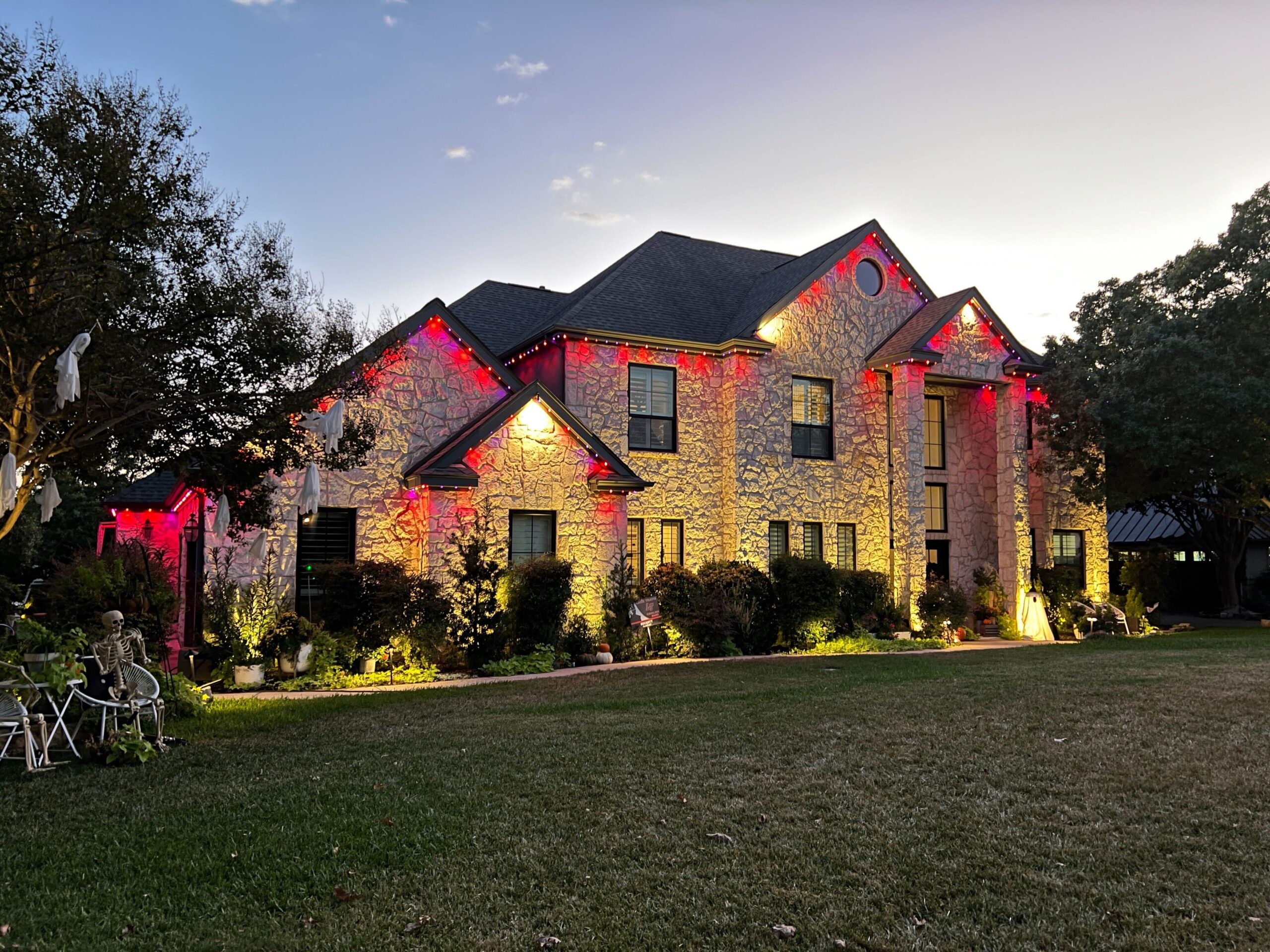 A two-story stone house illuminated with Astoria Permanent Outdoor Lights. The lights are set to a red color, lining the roof and highlighting the architectural details of the house.