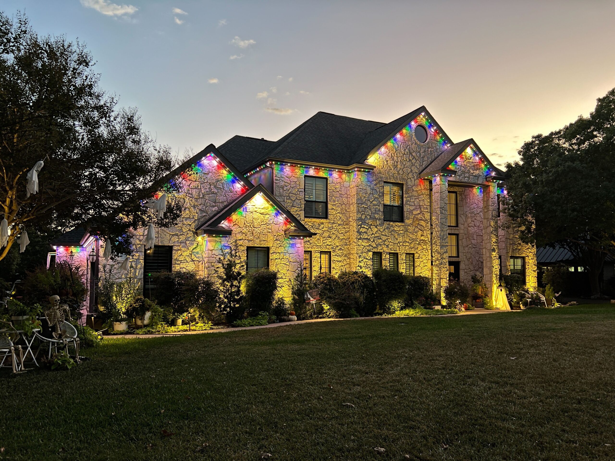 A two-story stone house illuminated with Astoria Permanent Outdoor Lights. The lights are set to a multicolor, lining the roof and highlighting the architectural details of the house.