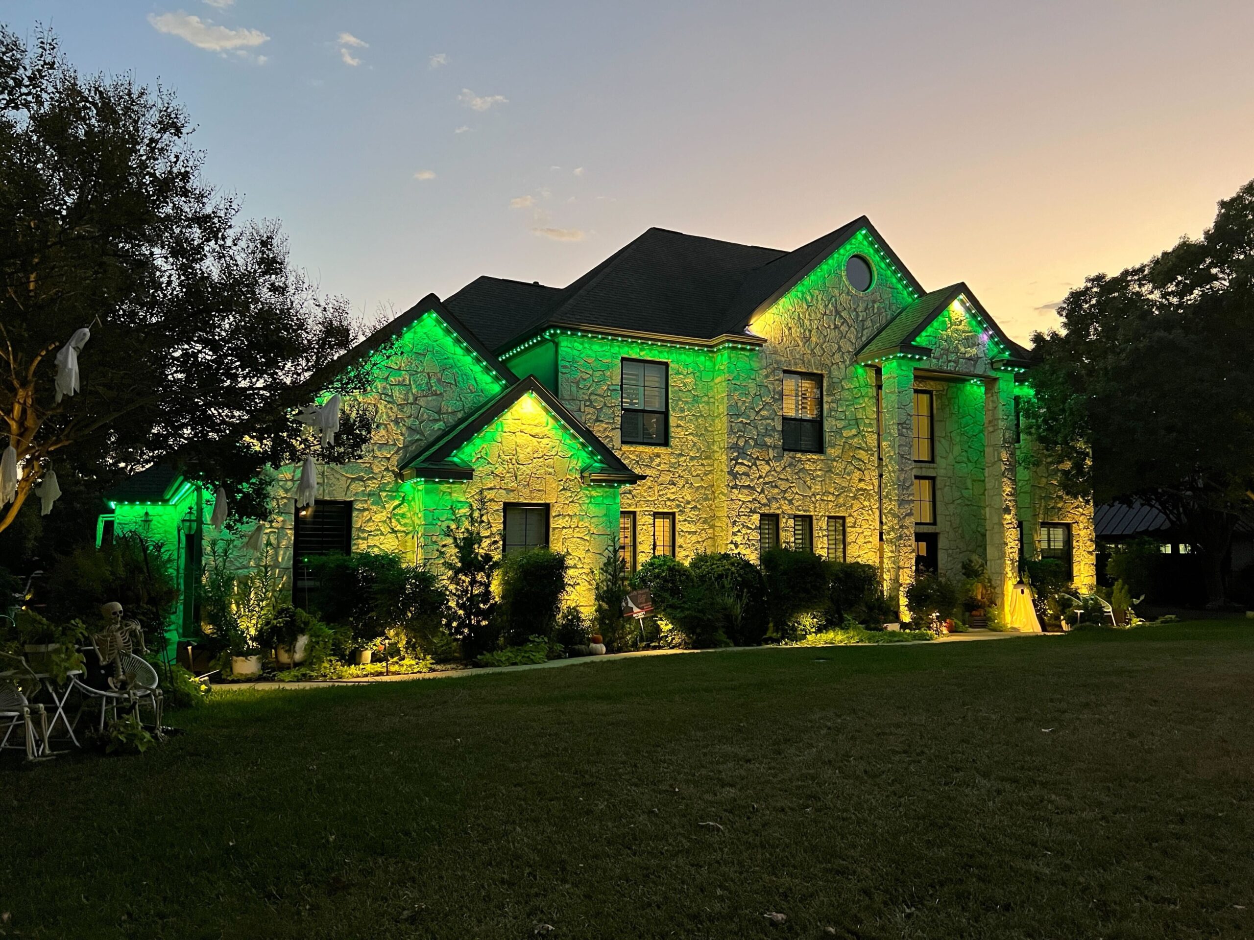 A two-story stone house illuminated with Astoria Permanent Outdoor Lights. The lights are set to a green color, lining the roof and highlighting the architectural details of the house.