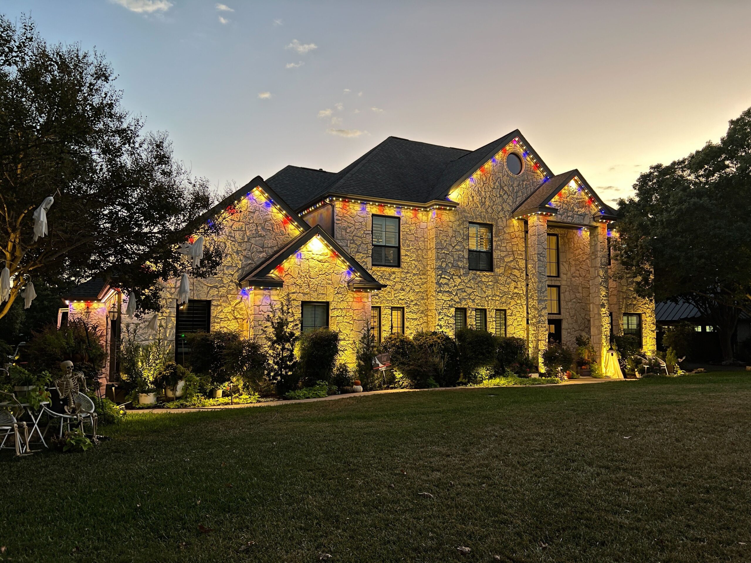 A two-story stone house illuminated with Astoria Permanent Outdoor Lights. The lights are set to a yellow, blue and red color, lining the roof and highlighting the architectural details of the house.