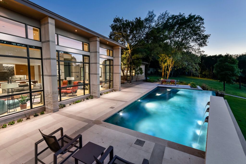 exterior of a modern house at dusk, featuring a swimming pool and patio area. The pool is lit from within. Trees and landscaping surround the property, and soffit lighting is visible on the house's exterior.