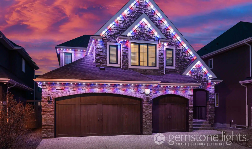 A two-story stone house illuminated with Gemstone Lights at dusk. The lights are installed along the rooflines, outlining the shape of the house with a mix of red, white, and blue colors.