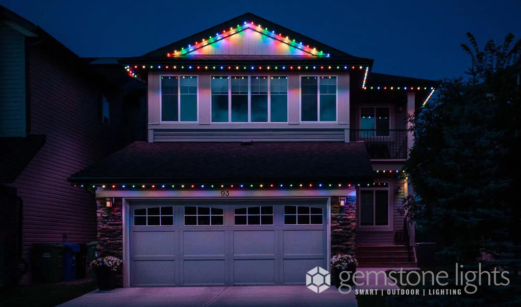 A modern two-story home with a garage, illuminated by vibrant permanent holiday lights in a multicolor pattern along the roofline.