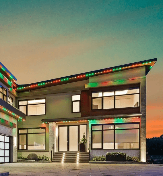 A modern luxury home, featuring permanent holiday lights in red and green along the roofline and architectural edges.