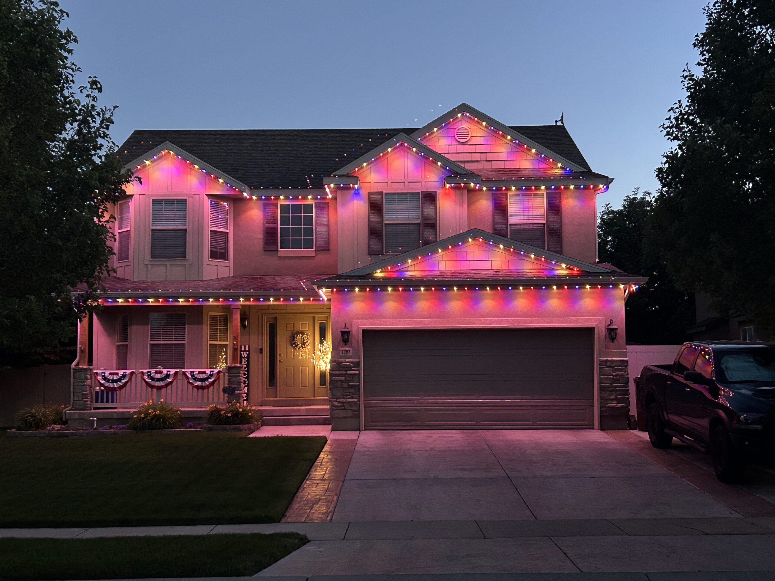 a two-story house decorated with multicolored Permanent Holiday Lighting at dusk. The lights outline the roofline and porch, and patriotic bunting hangs on the porch.