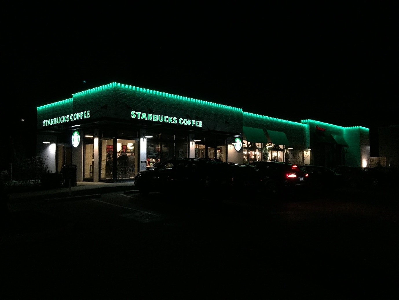 A Starbucks coffee shop warmly lit at night with vibrant green permanent holiday lights outlining the roofline and windows.