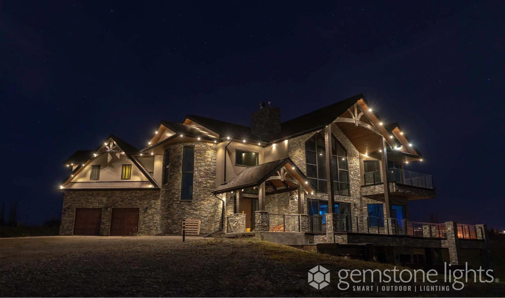 A large rustic home with a stone and wood exterior, illuminated by permanent warm white holiday lights along the roofline and architectural accents at night.