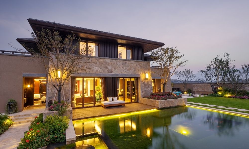 A house with warm lighting enhances the ambiance, with lights illuminating the deck, the house's exterior, and potentially the pool area.