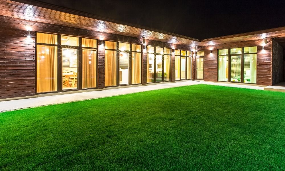 A modern house with large glass windows and wooden paneling, illuminated by warm exterior wall lights.