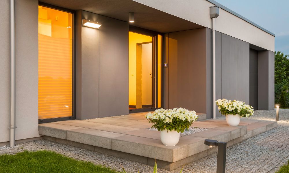 A modern house entrance with an outdoor wall and pathway lights illuminate the entrance and the surrounding gravel walkway.