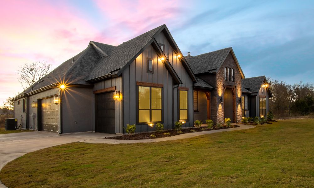 A gray modern farmhouse with a mix of vertical and brick siding, illuminated by warm outdoor lights, set against a twilight sky