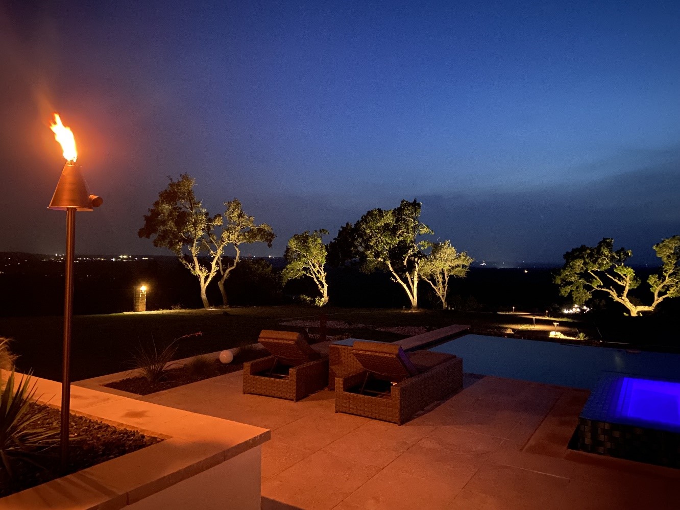 Luxurious outdoor pool area at night, surrounded by lush trees. Warm lighting softly illuminates the trees, casting reflections on the water. Lounge chairs and a cozy seating area create an inviting ambiance.