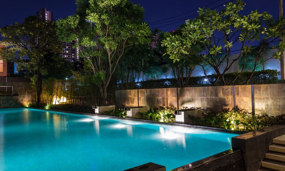 A beautifully lit outdoor swimming pool at night, surrounded by lush greenery and modern landscape lighting. The ambient glow reflects off the water, creating a serene and luxurious atmosphere.