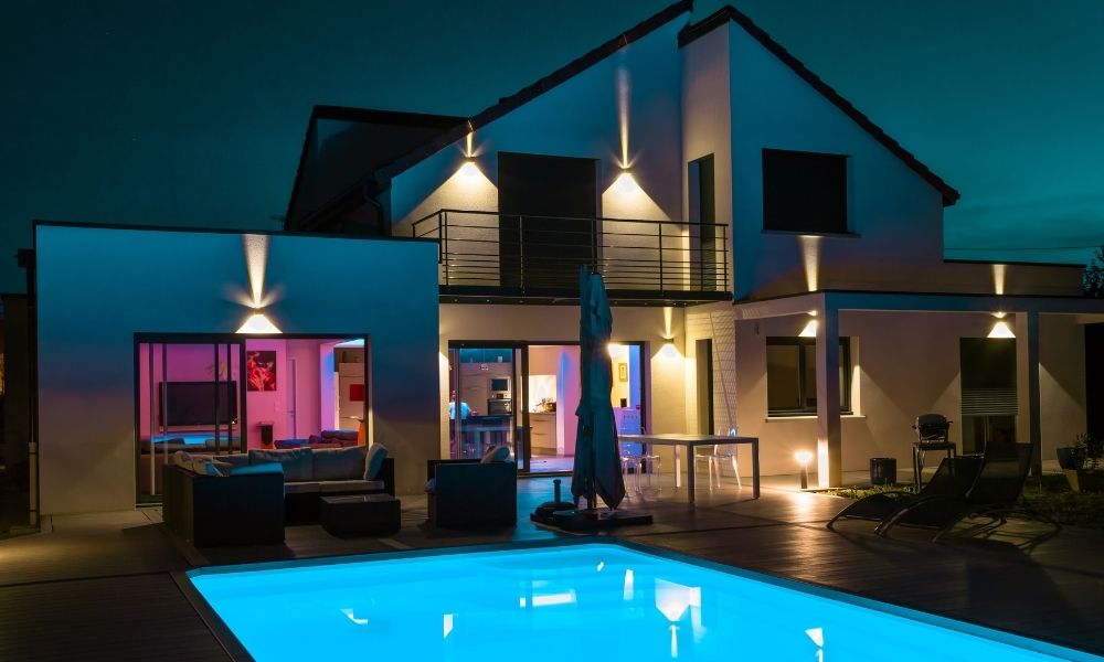 A modern house at night featuring elegant outdoor lighting and a beautifully illuminated swimming pool. The warm exterior lights highlight the architecture, while the blue glow of the pool enhances the contemporary ambiance.