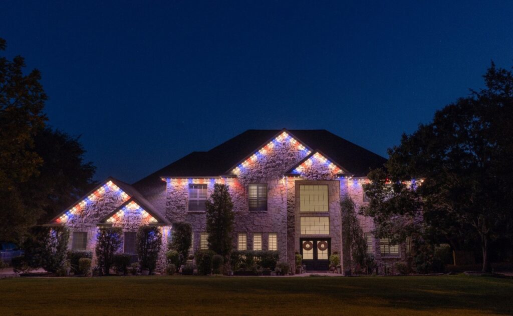 A luxurious brick and stone home is beautifully illuminated at night by blue red and white Astoria Lighting Co Lights. The elegant lighting highlights the architectural details.