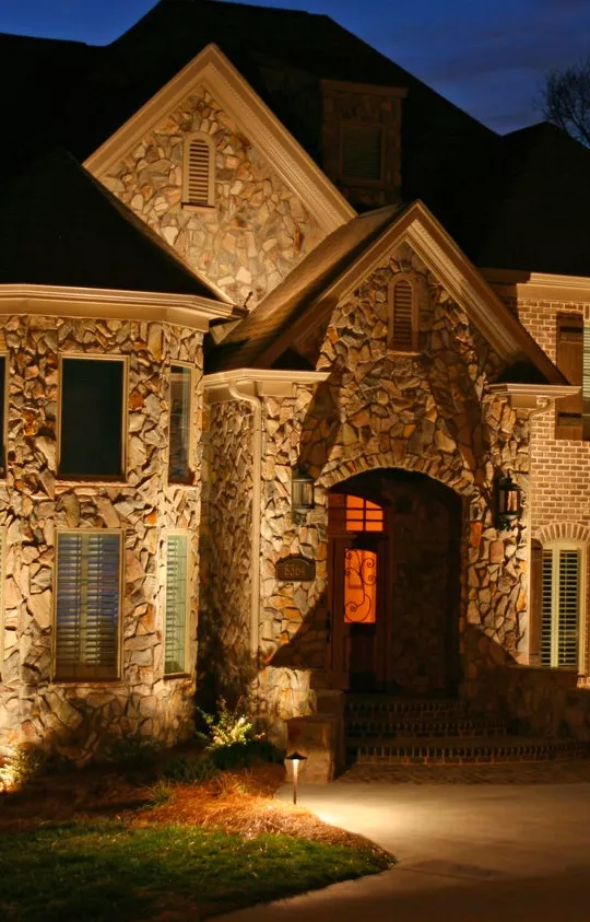 exterior of a stone house at night, illuminated by landscape lighting. The warm glow of the lighting highlights the stonework and creates a welcoming ambiance.