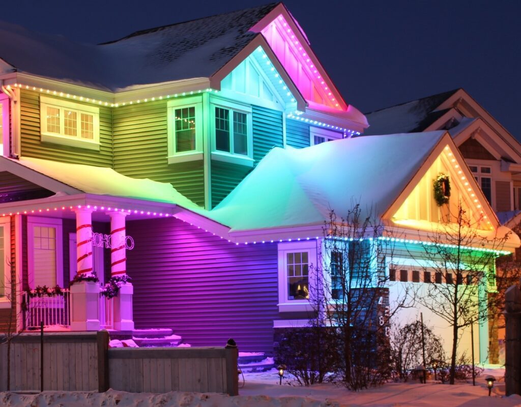 A modern and cozy house covered in snow. Permanent holiday lights in vibrant colors illuminate the roofline, adding festive charm. Exterior Architectural (Accent) Lighting enhances the home's sleek design.