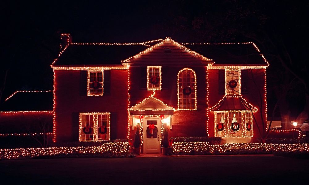 A two-story brick house with warm white and red Christmas lights outlining the roofline, windows, and entrance.