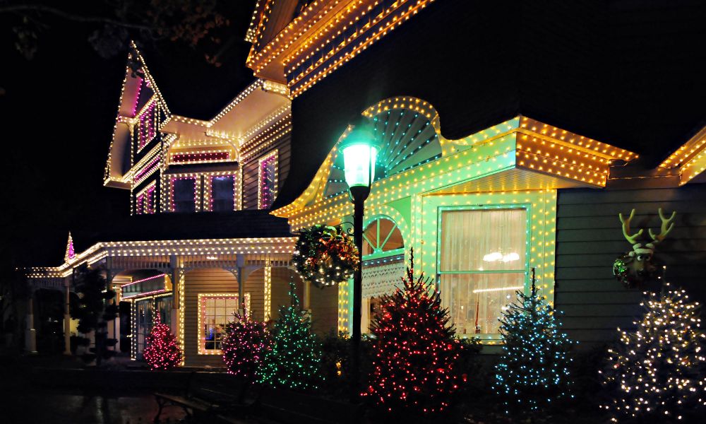 Warm permanent holiday lights elegantly installed along the roofline and walls of a house, creating a cozy and festive ambiance. The bushes in the front yard are decorated with Christmas lights in various colors.