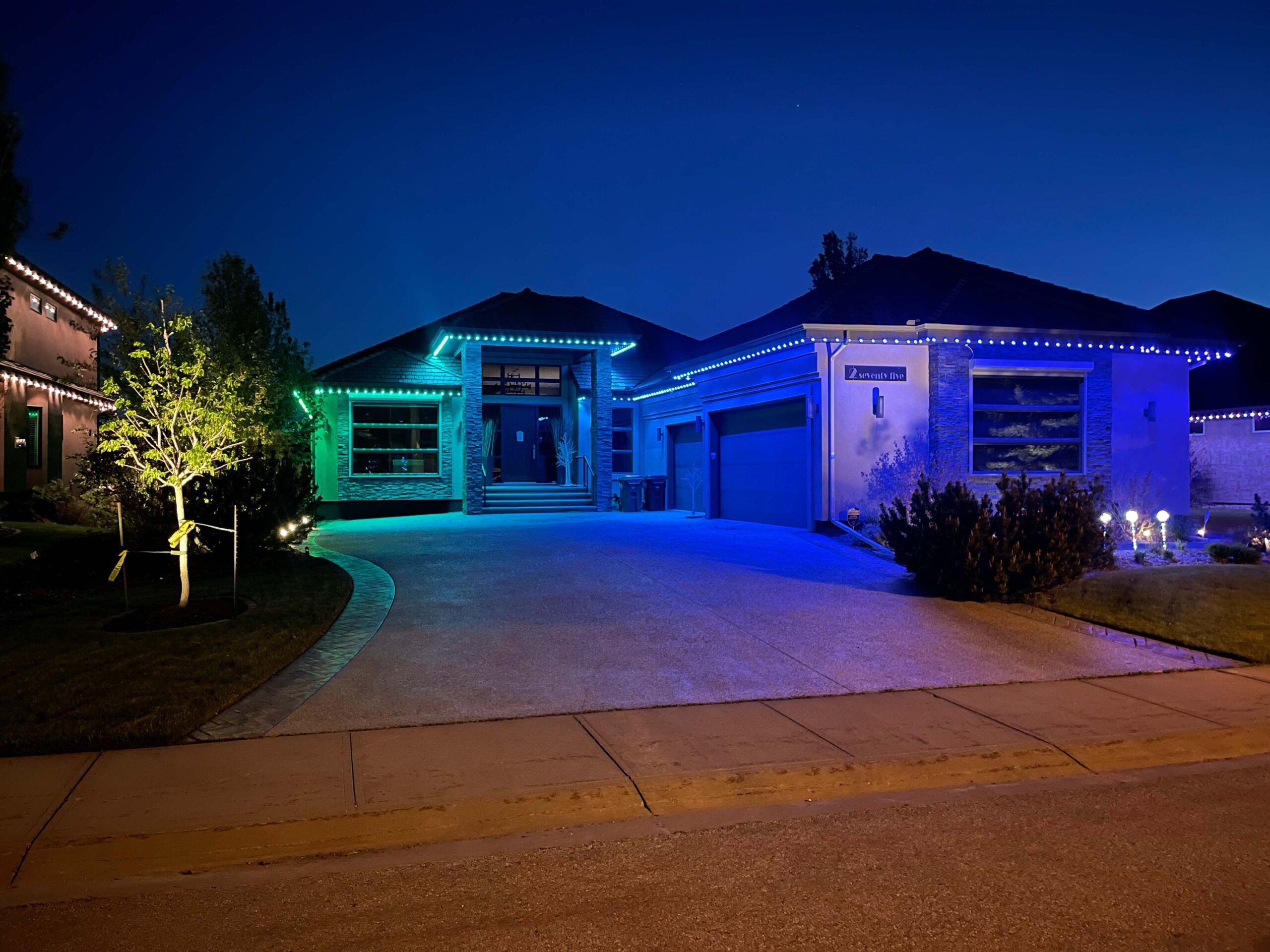 Residential home with driveway and garage featuring Astoria’s soffit-fitted permanent lighting system displaying a green and blue gradient pattern that enhances curb appeal and modern style.