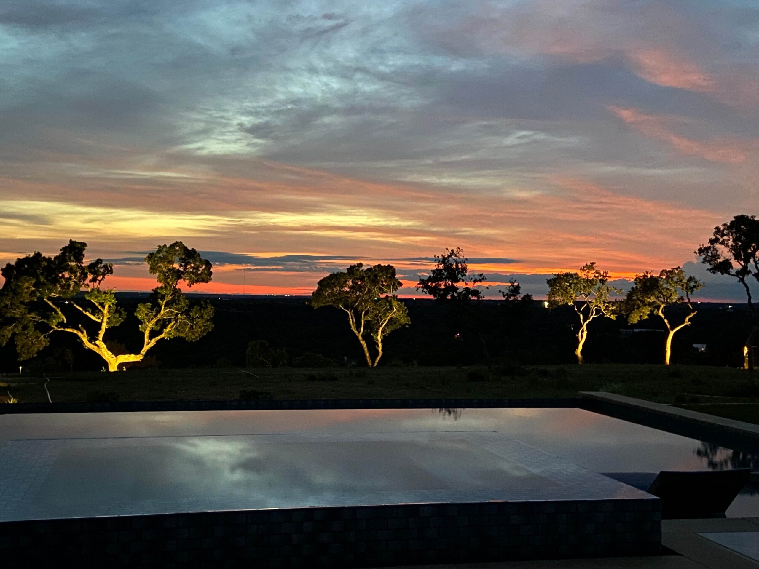 Luxury backyard featuring a pool with skyline views and Astoria’s landscape uplighting, highlighting the natural structure of mature trees and enhancing the property’s architectural elegance.