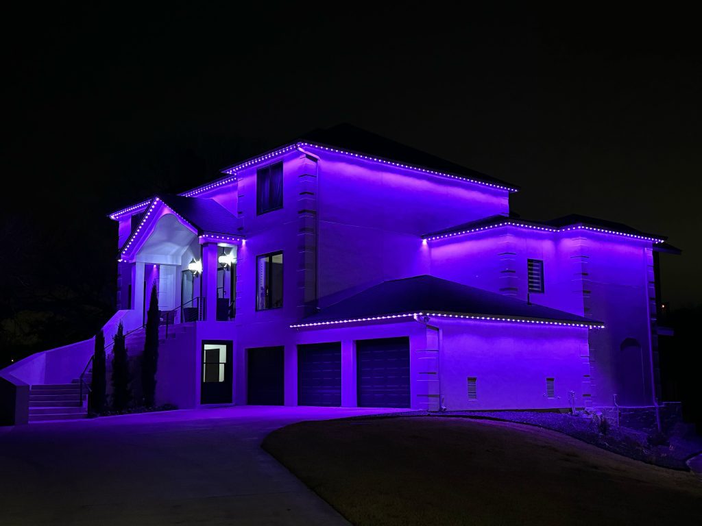 Residential white exterior home featuring Astoria’s permanent lighting system installed along the soffit line, displaying a vibrant purple glow against the night sky.