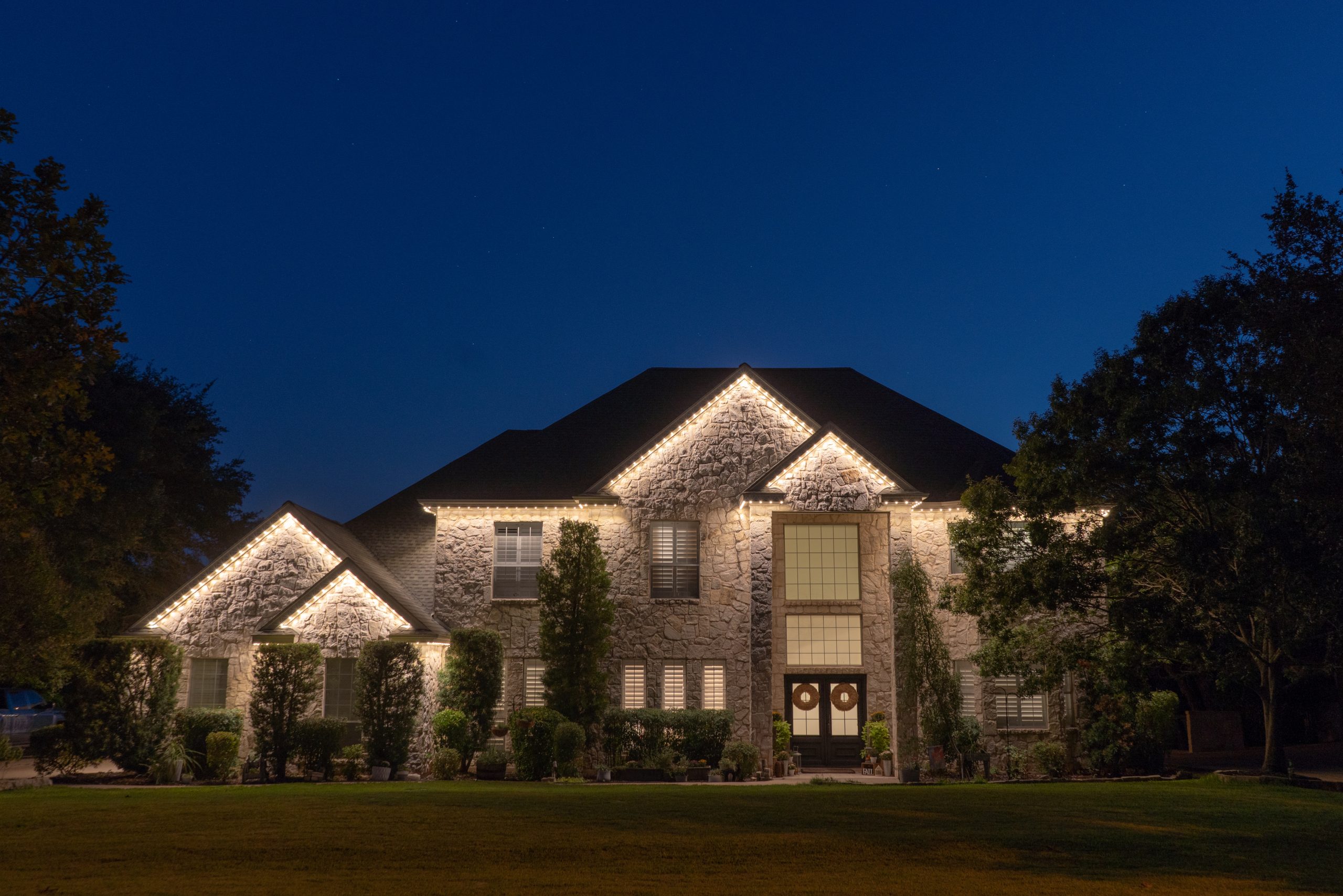 Luxury residential home with stone exterior and soffit line fitted with Astoria’s permanent holiday lighting system, displaying warm white lights that illuminate the architecture against a clear night sky.