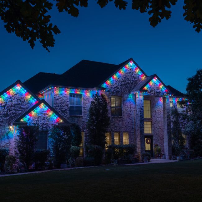 Luxury organic stone house featuring Astoria permanent lighting system along soffit line displaying rainbow pattern theme light for Easter Holiday.