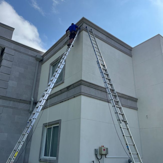Astoria installer adjusting permanent lighting along the soffit line of a luxury two-story home, viewed from a side angle in daylight.