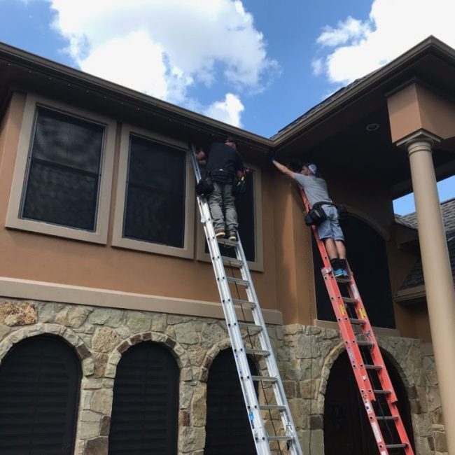 Astoria installers securing the closed-track lighting system along a two-story home’s soffit line using ladders and hand tools.